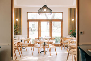 Sunlit modern restaurant interior with round wooden tables and chairs, large wood-framed windows, a woven pendant light, potted plants and minimalist décor.