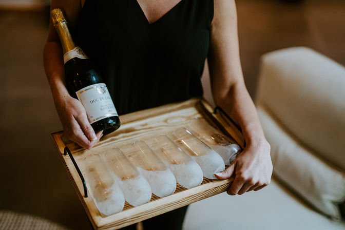 Server in a black dress holding a wooden tray with six frosted chilled champagne flutes and a bottle of sparkling wine, ready for an indoor reception.
