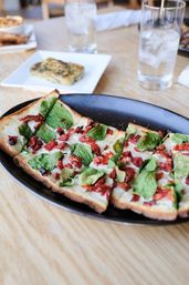 Sliced rectangular flatbread pizza with melted mozzarella, fresh spinach and sun-dried tomatoes on a black platter, served on a light wood table with glasses of iced water.