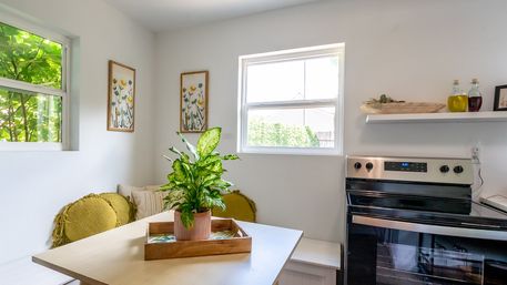 Sunlit cozy kitchen nook with wooden breakfast table, potted green houseplant in a tray, yellow round cushions, floral wall art, and a stainless steel stove under a bright window.