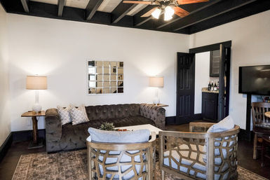 Cozy modern-rustic living room with dark exposed ceiling beams, tufted gray sofa, two lattice wooden accent chairs around a coffee table, mirrored wall art, table lamps and ceiling fan lighting.