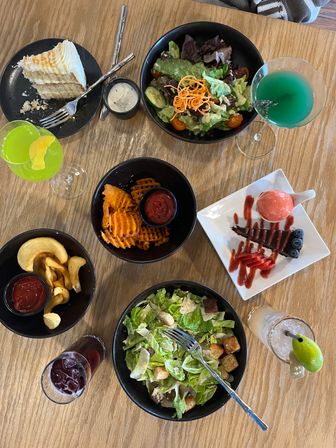 Overhead shot of a wooden restaurant table with two salads, waffle fries and potato curls with ketchup, a slice of layered cake, chocolate tart with sorbet and berries, and bright green and blue cocktails.