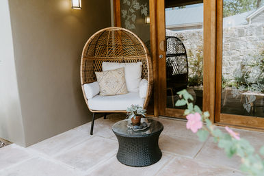 Cozy covered patio with a wicker egg chair upholstered in white and a patterned throw pillow, round black wicker side table with a potted succulent, sliding wood-and-glass doors and stone tile floor.