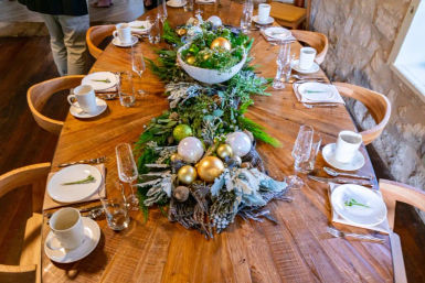 Rustic wooden dining table set for a holiday meal with lush green garland and gold and white baubles centerpiece, white plates, cups and glassware in a stone-walled room.