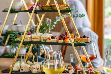 Elegant three-tier gold stand with assorted finger sandwiches, fruit-topped canapés and mini tarts, surrounded by wine glasses on a brunch table