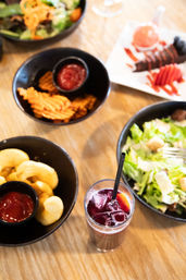 Top-down view of a casual restaurant table on wood: a tall iced berry drink with straw, bowls of green salad, waffle fries and potato puffs with ketchup, and a drizzled dessert.