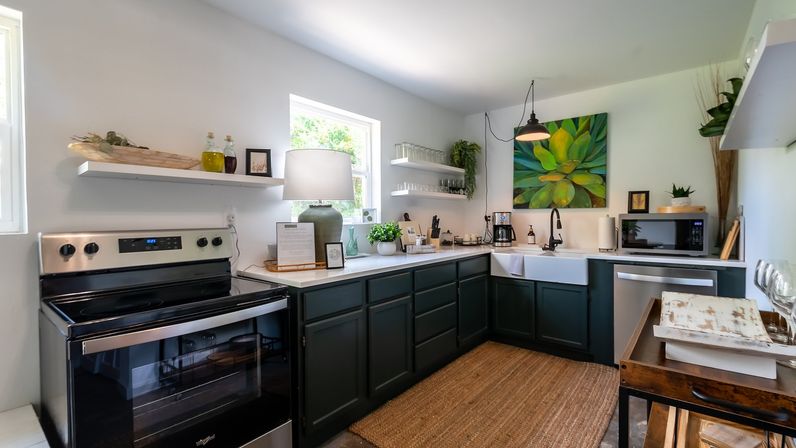 Sunlit modern farmhouse kitchen with dark green lower cabinets, white countertops, stainless steel oven, farmhouse sink, open shelves, potted plants and succulent wall art.