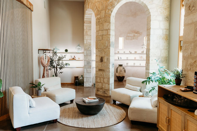 Sunlit, cozy lounge with exposed stone arches, four cream upholstered chairs around a round dark wood coffee table on a woven rug, potted plants, minimalist shelving with decorative vases and soft natural light