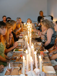 Candlelit communal dinner at a long wooden table, rows of taper candles glowing as guests share plates, drinks and conversation in a cozy indoor gathering.