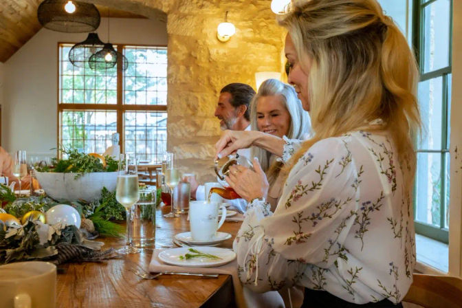 Sunlit rustic stone-walled dining room brunch — woman pouring tea into a white mug at a wooden table set with greenery centerpieces, champagne flutes and smiling friends nearby.