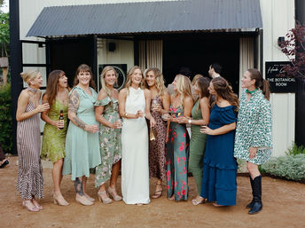 Bride in a white gown laughing with a group of bridesmaids and friends in colorful floral and patterned dresses, holding drinks outside a rustic barn-style wedding venue.