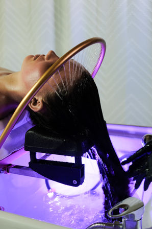 Client reclining at a salon hair-wash station, water arcing from a gold circular shower onto wet hair over a purple-lit backwash basin during a shampoo treatment.