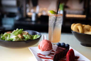 Restaurant table spread with a pink strawberry sorbet, sliced chocolate cake drizzled with berry sauce and topped with blueberries and a strawberry, paired with a Caesar salad, a bowl of onion rings and a tall lime cocktail with a straw.