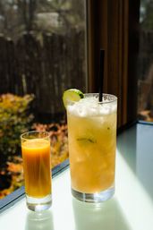 Sunlit patio table with a tall iced citrus cocktail garnished with a cucumber slice and black straw beside a small glass of bright mango juice