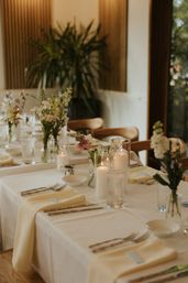 Candlelit, elegant dining table set for an intimate event with cream napkins, glassware, lit pillar candles and wildflower centerpieces by a sunlit window.