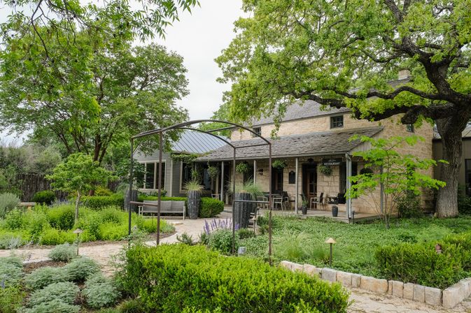 Stone farmhouse-style restaurant patio with hanging plants and wooden chairs overlooking a lush courtyard garden, stone paths and a large oak tree — peaceful country dining setting.