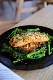 Close-up of cheesy herb-crusted chicken breast on stewed vegetables, surrounded by bright broccolini on a black plate over a rustic wooden table — restaurant-style healthy dinner.