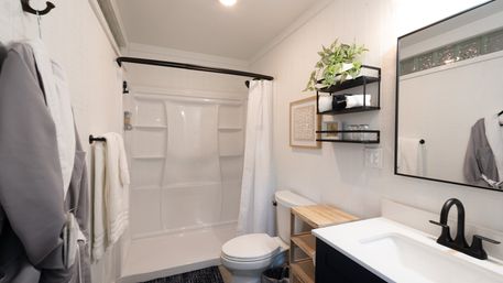 Bright modern small bathroom interior with white tub-shower, matte black fixtures and curved shower rod, wooden bench, wall shelves with plant and towels, and large rectangular mirror