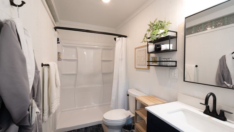 Bright modern small bathroom interior with white tub-shower, matte black fixtures and curved shower rod, wooden bench, wall shelves with plant and towels, and large rectangular mirror