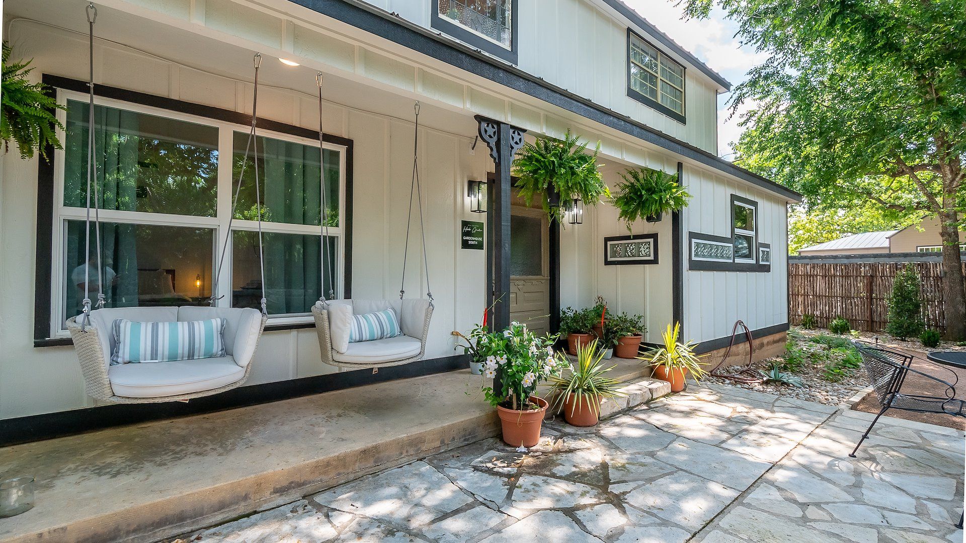 White farmhouse-style front porch with two hanging swing chairs, potted plants, and a stone patio in a shaded suburban yard.