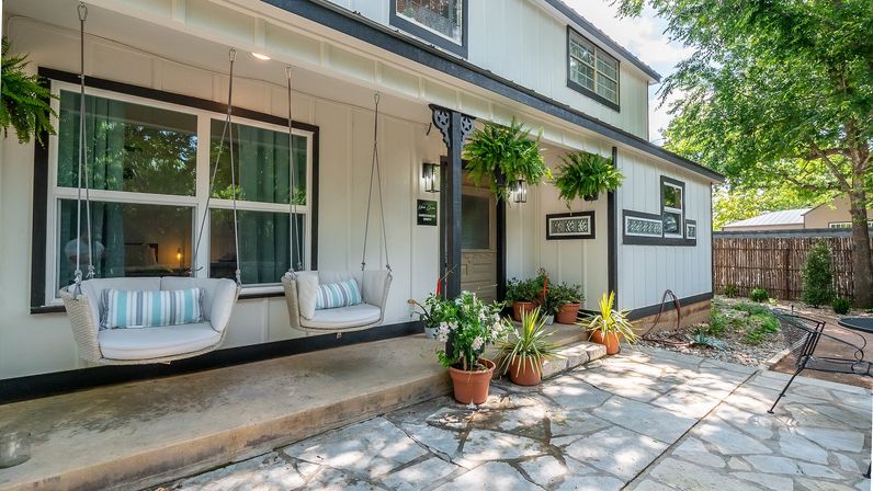 White farmhouse-style front porch with two hanging swing chairs, potted plants, and a stone patio in a shaded suburban yard.