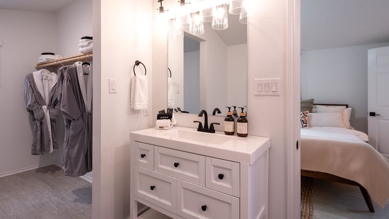 Bright modern master ensuite with a white vanity, black faucet and mirror lights, gray bathrobes hanging in an open closet, toiletry bottles on the sink, and a view into a cozy bedroom.