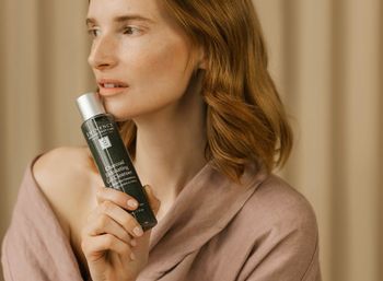 Woman holding a charcoal exfoliating gel cleanser bottle near her cheek in a soft, neutral-toned skincare lifestyle shot