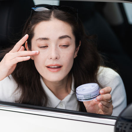 Woman applying face moisturizer from a jar while leaning out of a car window, sunglasses on her head and natural freckled skin — on-the-go skincare.