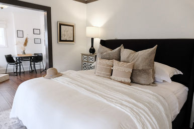 Bright neutral bedroom with white linens, layered textured throw and pillows, black upholstered headboard, bedside lamp and straw sun hat on the bed, hardwood floors and a small dining nook with round table and black chairs.