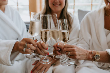 Three women in white robes clinking champagne flutes for a bridal-party toast in a sunlit getting-ready suite.