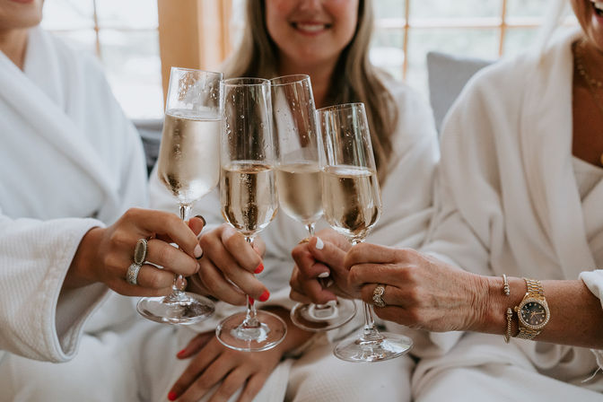 Three women in white robes clinking champagne flutes for a bridal-party toast in a sunlit getting-ready suite.
