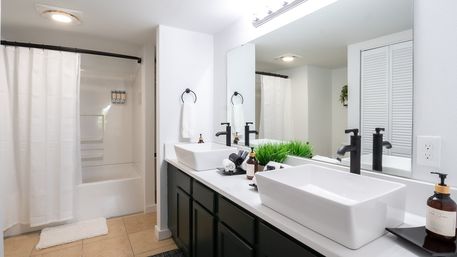 Bright modern bathroom interior with double white vessel sinks, matte-black faucets, large mirror, shower with white curtain, dark vanity cabinets and decorative potted grass