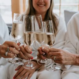 Close-up of four champagne flutes clinking in a sunlit living room toast, hands with rings and colorful nails.