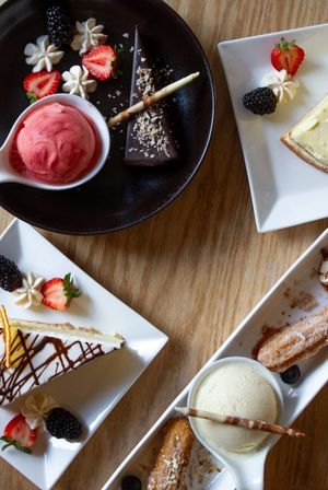 Overhead restaurant-style dessert spread on a wood table: scoop of strawberry sorbet with a chocolate tart, drizzled cheesecake slices with strawberries and blackberries, and vanilla ice cream with churros and a chocolate wafer stick.