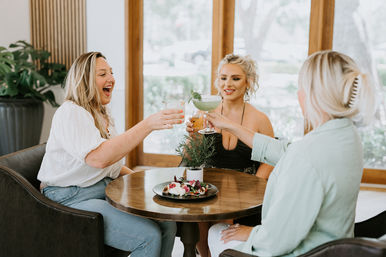 Three friends cheerfully toasting cocktails at a round wooden table in a bright café with large windows, a small potted herb and appetizer plate in the center — sunny brunch scene.