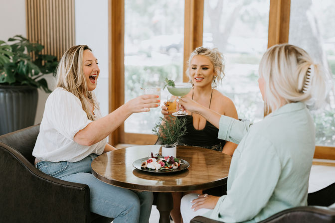 Three friends cheerfully toasting cocktails at a round wooden table in a bright café with large windows, a small potted herb and appetizer plate in the center — sunny brunch scene.