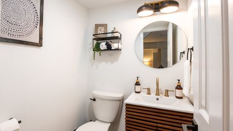 Bright modern powder room with white walls, round mirror, brass faucet and light fixture, wooden slatted vanity, toilet, and black wall shelf holding rolled towels and greenery.