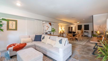 Cozy modern finished basement open-plan living and dining area with light-gray sectional sofa, wood dining table, area rug, indoor plants and warm lighting