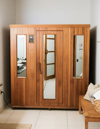 Cozy indoor wooden sauna cabin with glass windows and central door in a tiled home spa/bathroom corner, beside a wooden bench with towels and a woven basket.