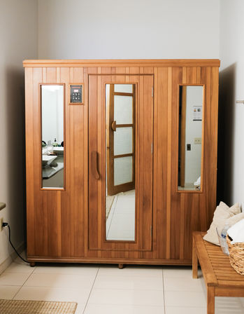 Cozy indoor wooden sauna cabin with glass windows and central door in a tiled home spa/bathroom corner, beside a wooden bench with towels and a woven basket.