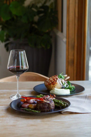 Fine-dining scene: glass of red wine and two plated entrees — seared steak with asparagus and roasted vegetables, and a grilled pork chop with mashed potatoes and greens on a wooden table by a window.
