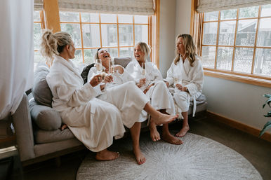 Four women in white robes laughing and clinking champagne glasses in a sunlit spa-style lounge with wooden-framed windows and neutral decor.