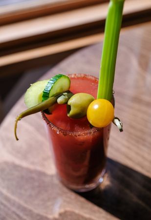 Spicy Bloody Mary in a tall glass on a wooden table, garnished with a celery stalk, cucumber slice, green olive, pepperoncini and a yellow cherry tomato with a chili-salt rim