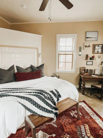 Cozy farmhouse-style bedroom interior with white paneled headboard, king bed in white linens, black-and-white striped throw and dark gray and burgundy pillows, wooden bench at the foot, red patterned area rug, wooden desk by a sunlit window, beige walls and ceiling fan.