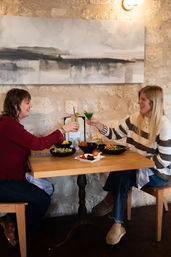 Two friends clinking cocktails over salads and desserts at a cozy stone‑walled bistro table beneath abstract wall art