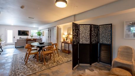Modern open-plan basement living and dining area with round wooden table and chairs on a patterned rug, gray sofa and wall TV, decorative black folding screen, wicker chair and console lamp