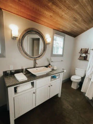 Cozy modern farmhouse bathroom with sloped wood-plank ceiling, round rustic mirror above a vessel sink on granite countertop, white vanity, towel shelves and toilet