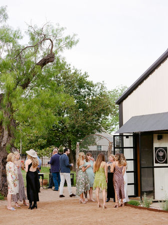 Outdoor summer gathering at a rustic barn-style venue — guests in colorful dresses mingle on a dirt courtyard under large trees beside a light metal building.