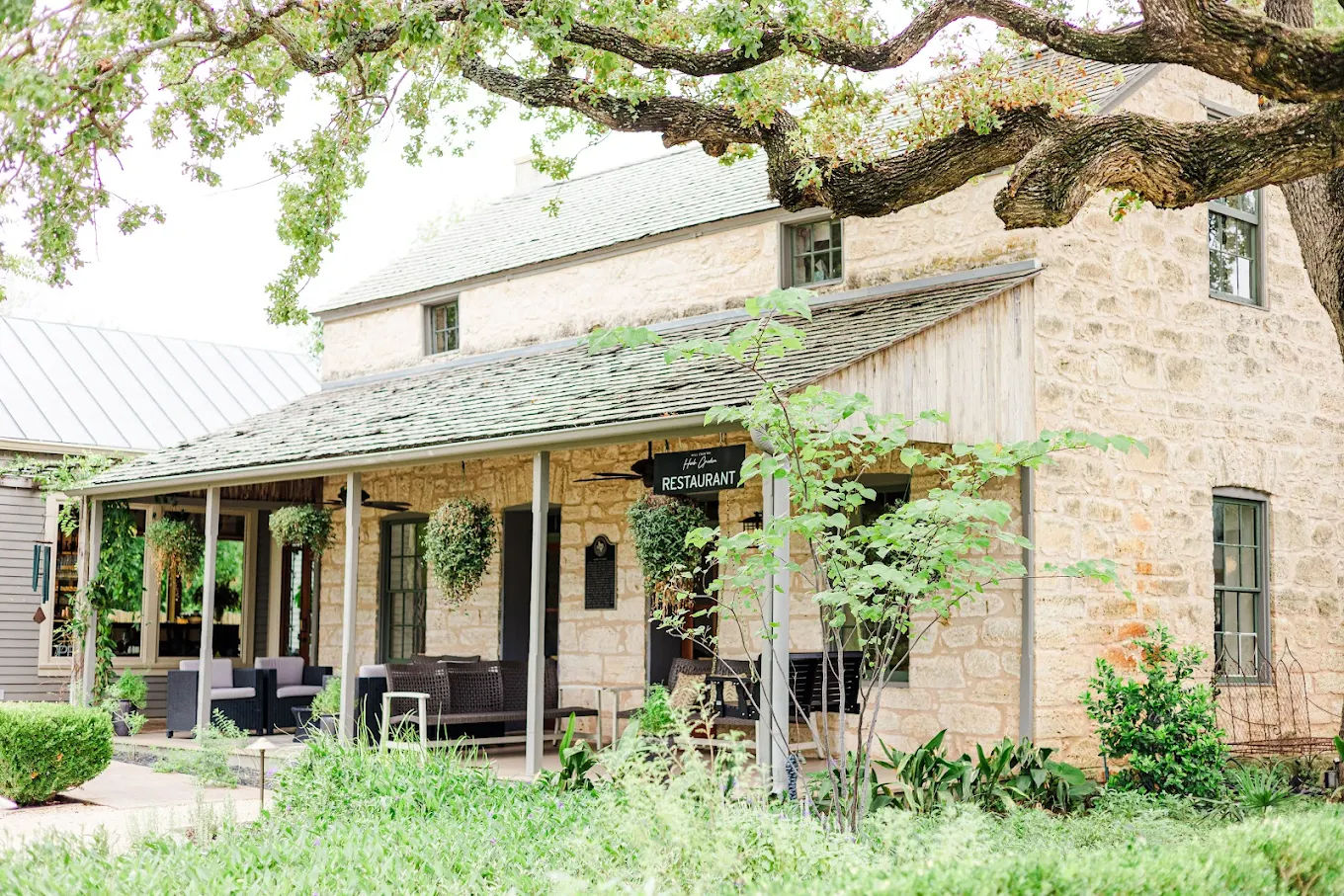 Rustic stone country restaurant with covered porch, hanging planters, outdoor seating and lush garden under large oak tree branches