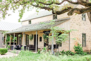 Rustic stone country restaurant with covered porch, hanging planters, outdoor seating and lush garden under large oak tree branches